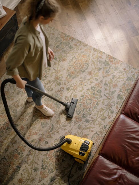 A person cleaning a carpet using a yellow and black handheld wet/dry vacuum cleaner, positioned on a patterned carpet in a residential living room. The individual's hands are shown inserting a cloth i